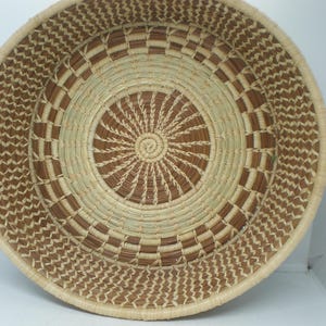 May include: A round, shallow woven basket featuring a coiled construction with alternating light beige and dark brown natural fibers. The design displays a central sunburst pattern surrounded by concentric rings of geometric block motifs.