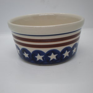 May include: Cream-colored ceramic bowl with a patriotic design. It features horizontal bands of blue and brown, and a row of blue circles with white stars. A classic piece for serving.
