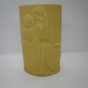 May include: A yellow ceramic vase with a raised design of a woman holding a glass. The vase is a rounded rectangle shape.