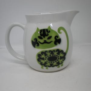 May include: White ceramic pitcher with a green and black cat design. The cat has a stylized face and body. The pitcher has a handle and a spout for pouring. A decorative kitchenware item.