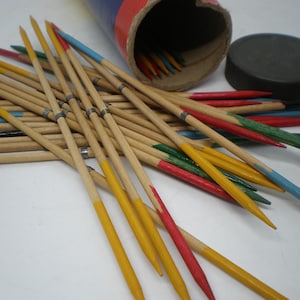 May include: A set of wooden pick-up sticks featuring pointed ends painted in yellow, red, green, and blue. The sticks are scattered in a pile with a cylindrical cardboard storage tube and a black plastic lid positioned in the background.