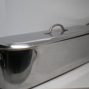 May include: A rectangular stainless steel fish poacher with a matching lid and two side handles. The polished metal surface reflects light, and the lid features a central arched handle for easy removal from the elongated cooking vessel.