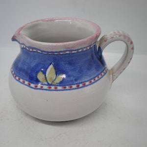 May include: A small, round ceramic pitcher with a handle and spout. The pitcher is white with a blue band around the middle and a pink rim. The blue band has a floral design and red dots.