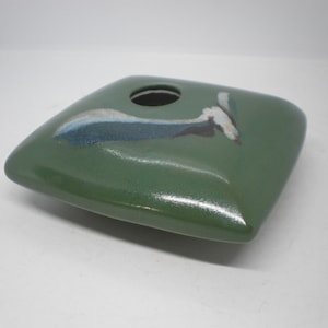 May include: A square, low-profile ceramic vase with a smooth, glossy forest green glaze. The top surface features a centered circular opening and an abstract, hand-painted brushstroke design in shades of white, dark blue, and black.