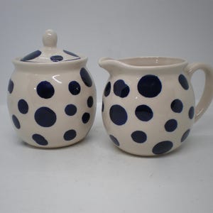 May include: Cream-colored ceramic sugar bowl with lid and creamer, both adorned with navy blue polka dots. The sugar bowl is round with a small knob on the lid. The creamer has a handle and a spout.