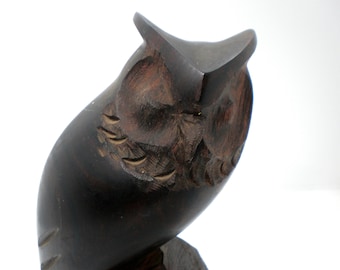 May include: A dark brown wooden owl figurine featuring hand-carved details around the eyes and face. The sculpture displays a smooth, polished finish with visible wood grain patterns and carved feather textures along the sides of the body.