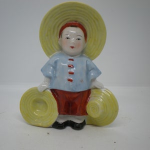 May include: A ceramic figurine of a person wearing a blue shirt, red pants, and a yellow straw hat. The person is holding two more yellow straw hats.