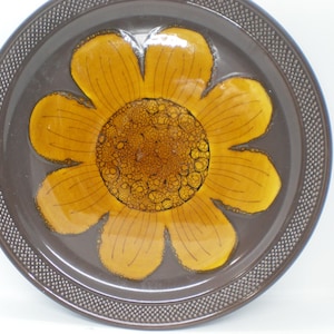 May include: A round ceramic plate featuring a large, central yellow flower design with a textured, dark brown center. The background of the plate is a deep brown, and the outer rim is decorated with a raised, dotted grid pattern in a matching brown.