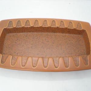 May include: A rectangular, brown ashtray with a speckled interior and a raised, notched rim. The ashtray has a rounded rectangular shape and is made of ceramic or a similar material. The notches are evenly spaced around the perimeter.