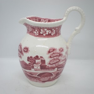 May include: A cream-colored ceramic pitcher featuring a deep red transferware pattern. The design depicts a landscape with a stone bridge, castle-like structures, and trees. A floral border encircles the rim, and a curved handle is attached to the side.