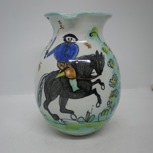 May include: White ceramic pitcher with a blue and yellow painted design of a person riding a black horse. The pitcher has a green and white floral design around the base.