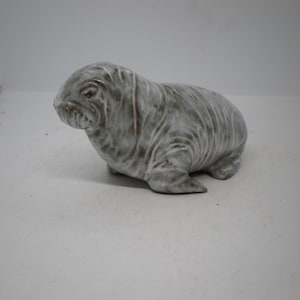 May include: A ceramic walrus figurine featuring a glossy, mottled grey and white glaze. The sculpture depicts the animal in a resting position with visible skin folds, a rounded head, and small flippers, showcasing a smooth, reflective surface.