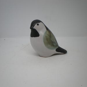 May include: A ceramic bird figurine featuring a smooth white body, a black head and throat patch, and a soft olive-green wing detail. The bird has a small black beak and a dark tail, presented in a rounded, stylized form with a matte finish.