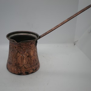 May include: A copper coffee pot with a long handle and an intricate engraved design. The pot is made of a dark copper color and has a traditional style.