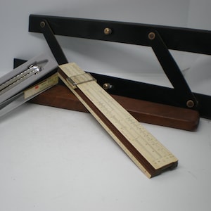 May include: A beige slide rule with a sliding metal cursor, a silver metal drafting compass in a gray plastic case, and a black articulated drafting arm with brass rivets. The items are arranged together, showcasing vintage technical drawing tools.