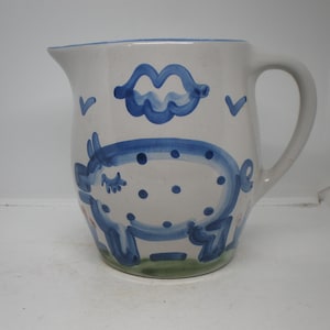 May include: White ceramic pitcher with a blue pig illustration. The pitcher has a curved handle and a spout for pouring. The pig is outlined in blue with blue polka dots. Blue clouds and birds are painted on the pitcher.