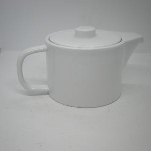 May include: A white ceramic teapot with a matching lid. The teapot has a curved handle and a spout for pouring. The design is simple and modern, suitable for serving tea.