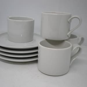 May include: A set of white ceramic mugs and saucers. There are four mugs with handles and a stack of five saucers. The mugs are stacked on top of each other and on the saucers.