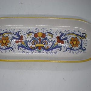 May include: A rectangular ceramic serving platter with a white base and a yellow rim. The platter features a central floral design in orange, blue, and yellow. The handles have red accents.