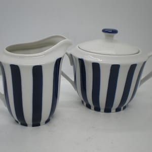 May include: A white creamer and sugar set with blue vertical stripes. The creamer has a handle and the sugar bowl has a lid.