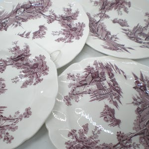 May include: Four white ceramic plates with a scalloped edge. Each plate features a detailed purple transferware design of a landscape with trees, buildings, and figures. The plates are arranged in a slightly overlapping pattern.