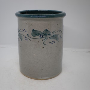 May include: A cylindrical stoneware utensil holder. The holder is light gray with a teal rim and a teal floral design. The design features leaves and small dots. The utensil holder is a kitchen decor item.