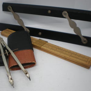 May include: A set of vintage drafting tools including a metal compass, a wooden folding ruler with black markings, and a black and brown leather case. Behind these items are two black parallel bars connected by two silver-toned metal linkages.