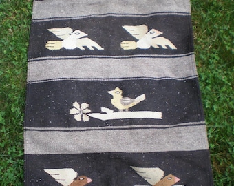 May include: A rectangular, woven textile with a repeating pattern of birds and floral elements. The background is black with horizontal gray stripes. The birds are depicted in shades of brown, beige, and white. The textile is likely a rug or wall hanging.