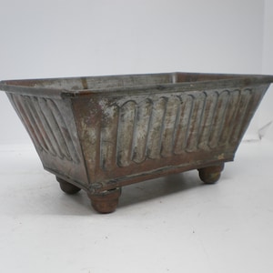 May include: A rectangular, aged metal planter with a weathered appearance. The planter features decorative vertical ridges on the sides and rests on four small, rounded feet. The overall color is a mix of brown and gray.