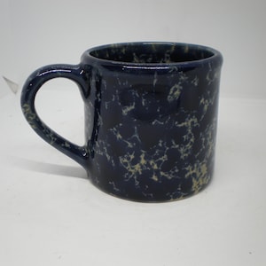 May include: A dark blue ceramic mug featuring a speckled, mottled pattern in light beige or off-white. The mug has a rounded handle on the left side and a cylindrical body with a smooth, glossy finish throughout the exterior surface.