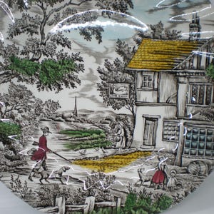 May include: A round ceramic plate featuring a detailed black and white transfer print of a rural English scene with a building labeled The Swan, a person walking a dog, and figures in the foreground, accented with touches of green, red, and yellow.
