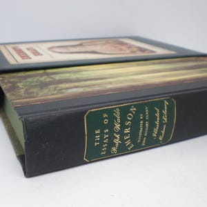 Pode incluir: Um livro preto de capa dura com letras douradas na lombada. O t&iacute;tulo &eacute; "The Essays of Ralph Waldo Emerson" com as palavras "Illustrated by John Stuart Curry" e "The Illustrated Library" abaixo do t&iacute;tulo.