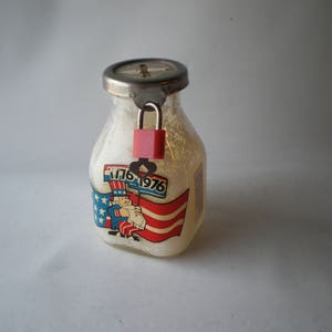 May include: A glass piggy bank shaped like a milk bottle with a metal lid and a padlock. The bottle has a label with a cartoon image of Uncle Sam holding a flag with the text "1776 1976".