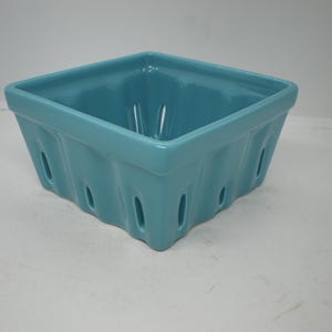 May include: A square, turquoise ceramic berry basket with a glossy finish. The basket has a series of oval-shaped openings along the sides, and a smooth, rounded rim. The design is reminiscent of traditional fruit containers.