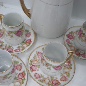 May include: A white porcelain tea set with a floral rose pattern. The set includes a teapot, four teacups, and saucers. The cups and saucers have gold trim. The roses are pink and cream colored.