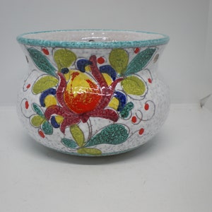 May include: White ceramic hanging planter with a floral design. The planter features a large red, yellow, and blue flower with green leaves. The planter has a speckled white glaze and a turquoise rim.