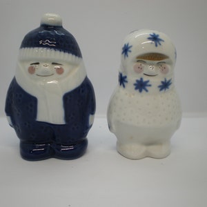 May include: A pair of ceramic salt and pepper shakers in the shape of people wearing winter clothing. The man is wearing a blue hat and coat, and the woman is wearing a white hat with blue stars. Both have rosy cheeks.