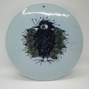 May include: A round ceramic plate with a blue and white glaze. A stylized bird with green, red, and yellow details is painted on the plate.
