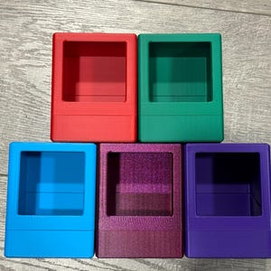 May include: Five colorful, square-shaped containers in red, green, blue, purple, and a glittery plum color. Each container has a rectangular opening at the top, suggesting they are designed for storage or display of small items.