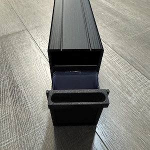 May include: A black, rectangular card holder with a pull-out drawer. The top of the holder has three vertical grooves. The drawer is open, revealing a stack of cards. The holder is made of a matte black material.