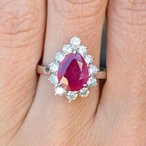 Può includere: Un anello d'argento con una grande pietra preziosa rosso scuro a forma di pera, circondata da un alone di pietre preziose più piccole e chiare. L'anello è posto su uno sfondo color carne.
