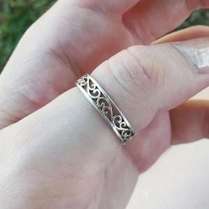 Peut inclure: Une bague argentée avec un motif ajouré orné de motifs tourbillonnants et symétriques. La bague est portée au doigt, mettant en valeur ses détails complexes et son savoir-faire. Le design de la bague est visible de tous les angles.