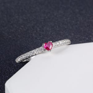 Peut inclure: Une bague en argent ornée d'une pierre précieuse rose vif en forme de cœur. La bande est ornée de petits pierres précieuses claires. La bague est posée sur un fond blanc et sombre.