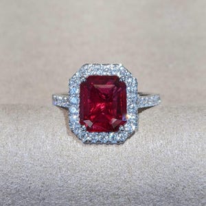 May include: A silver ring featuring a large, rectangular, deep red gemstone surrounded by a halo of smaller, clear stones. The band is also adorned with small, clear stones. The ring is set against a neutral background.