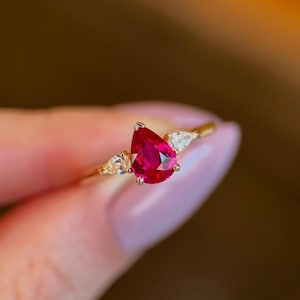 Può includere: Un anello d'oro con una gemma rossa a forma di pera, vibrante come elemento centrale, affiancata da due gemme più piccole e trasparenti. La fascia dell'anello è di un delicato colore oro, creando un design elegante e accattivante. È un gioiello.