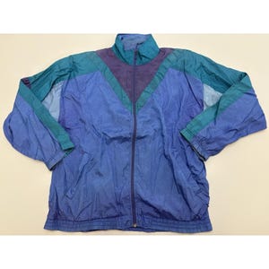 Könnte beinhalten: Vintage Trainingsjacke in Blau-, Türkis- und Lilatönen. Die Jacke hat einen durchgehenden Reißverschluss, einen hohen Kragen und ein farblich abgesetztes Design. Die Ärmel sind hellblau, und die Jacke hat einen gerippten Saum und Bündchen.