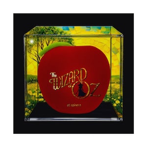 Op de afbeelding: Een rode appel met een groen blad in een doorzichtige vitrine. De appel heeft gouden tekst met de tekst "The Wizard of Oz" met een silhouet van een personage. De achtergrond is een geel en groen landschap.