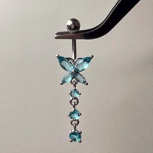 May include: A silver belly button ring featuring a butterfly design. The butterfly and dangling elements are embellished with light blue gemstones. The ring is held by tweezers against a plain backdrop.