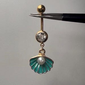 May include: A gold-coloured belly button ring with a dangling teal seashell charm. The ring features a clear gemstone and a small pearl within the shell. The top of the ring has a small gold ball.
