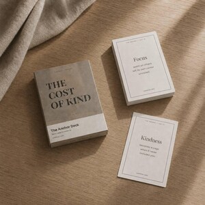 May include: A set of The Anchor Deck cards with a neutral color palette. The box reads "THE COST OF KIND" and the cards display the words "Focus" and "Kindness" with accompanying text. The cards are on a wooden surface.